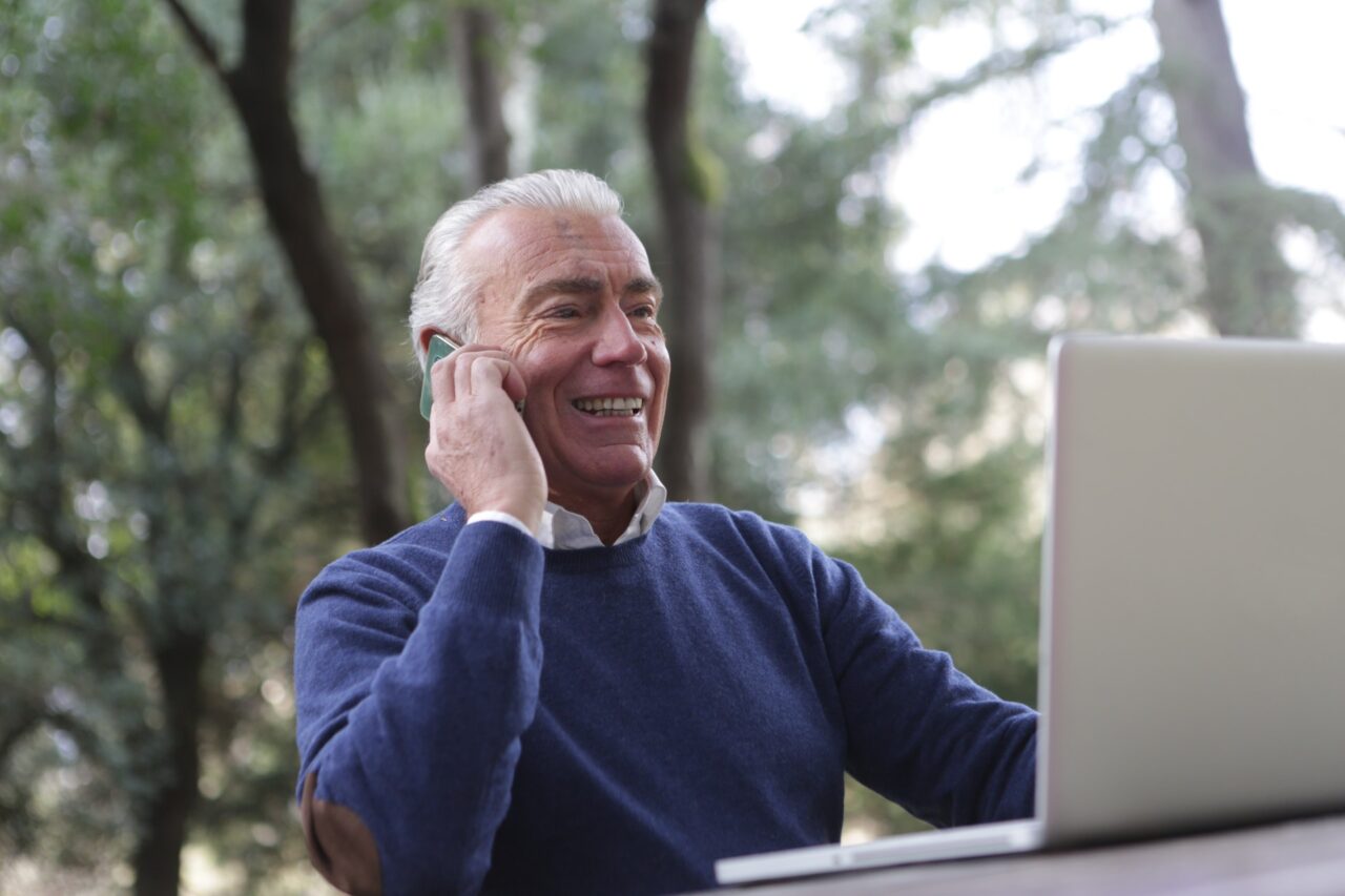 Older man smiling on the phone while looking at his laptop