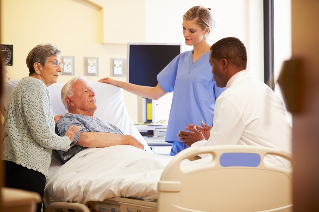 Picture of two medical professionals and an older woman conversing over an older man in a hospital bed