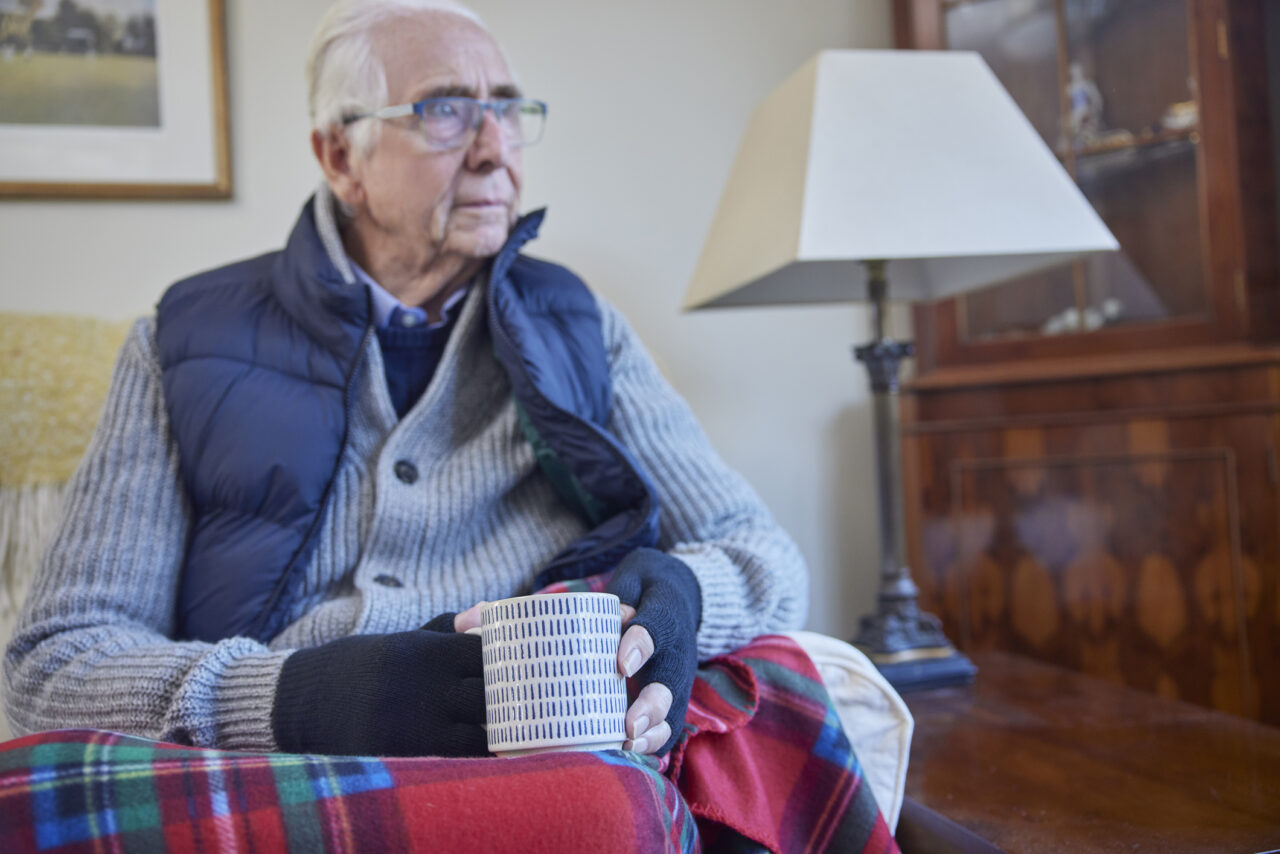 Image of older man Wearing Extra Clothes With Hot Drink Trying To Keep Warm At Home In Energy Crisis