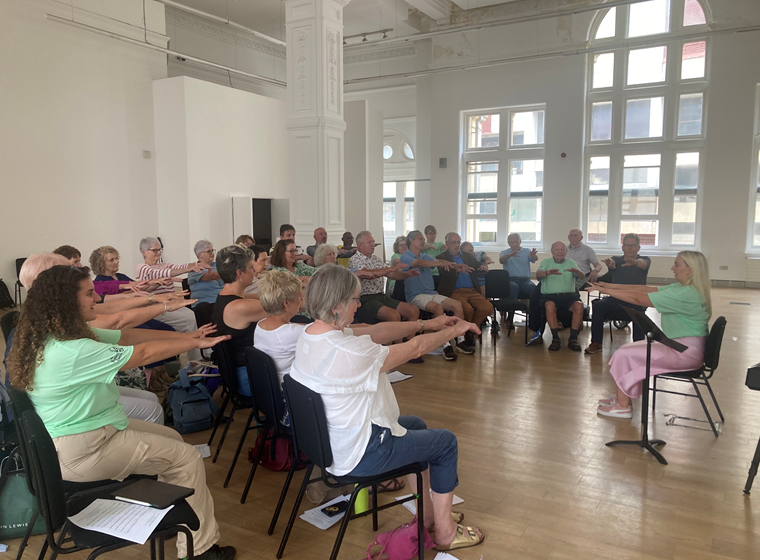 Delwedd o tua thri deg o bobl yn mynychu Côr Good Vibrations, yn eistedd ar gadeiriau mewn hanner cylch o amgylch arweinydd, yn dal eu breichiau allan o'u blaenau, yn ymarfer canu.