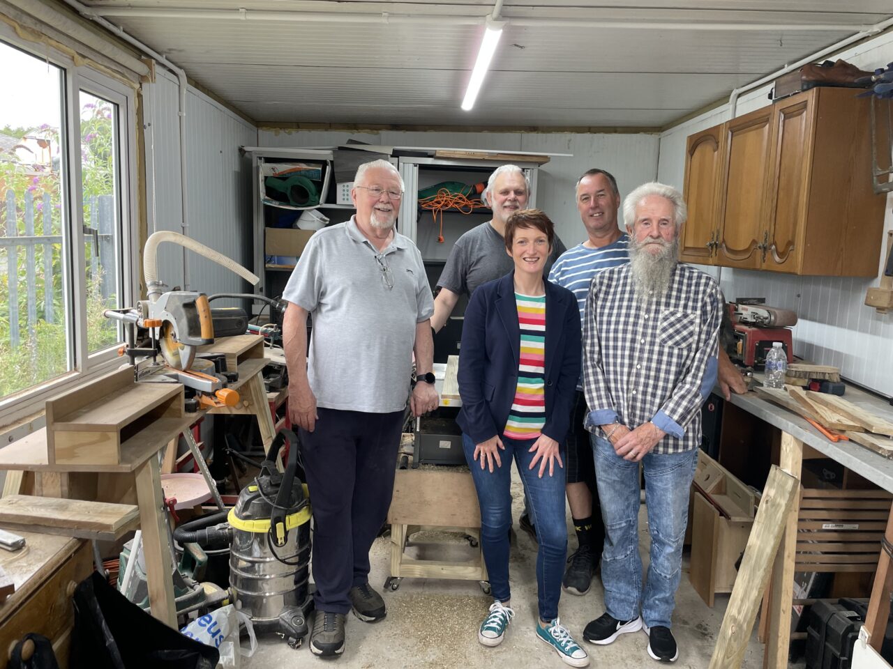 Delwedd o Gomisiynydd Pobl Hŷn Cymru, Rhian Bowen-Davies, yn y llun ochr yn ochr â phedwar aelod o The Shed yn Llansamlet. Mae'r pum person i gyd yn sefyll y tu mewn i sied adeiladu, o flaen turn.