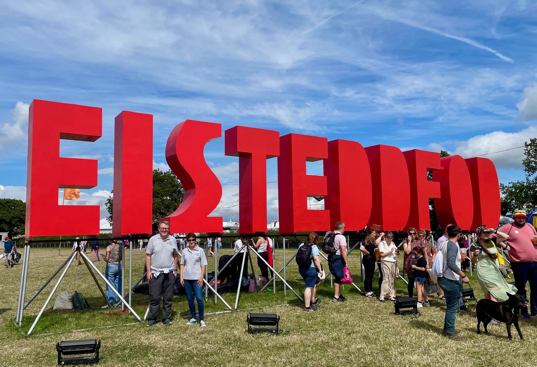 Delwedd o Rhian Bowen-Davies a'i chydweithiwr o flaen yr arwydd coch mawr 'EISTEDDFOD'.