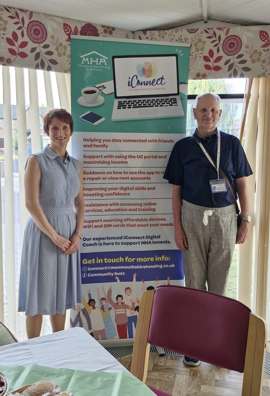 Comisiynydd Pobl Hŷn Cymru, Rhian Bowen-Davies, yn y llun ochr yn ochr ag Alistair, gwirfoddolwr o iConnect, yn sefyll o flaen baner yn hyrwyddo'r gwasanaeth iConnect.
