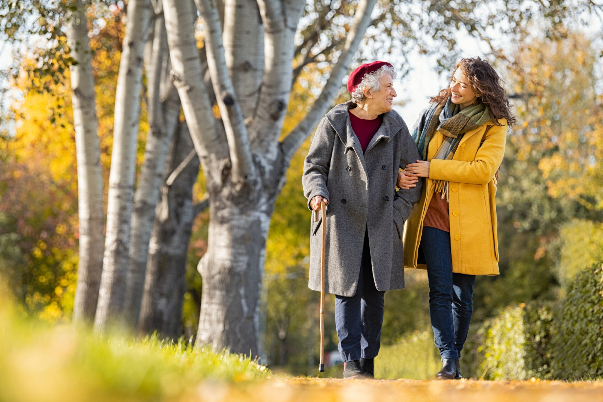 Delwedd o fenyw hŷn yn cerdded drwy'r parc, fraich ym mraich gyda gofalwr iau. // Image of an older woman walking through the park, arm in arm with a younger carer.