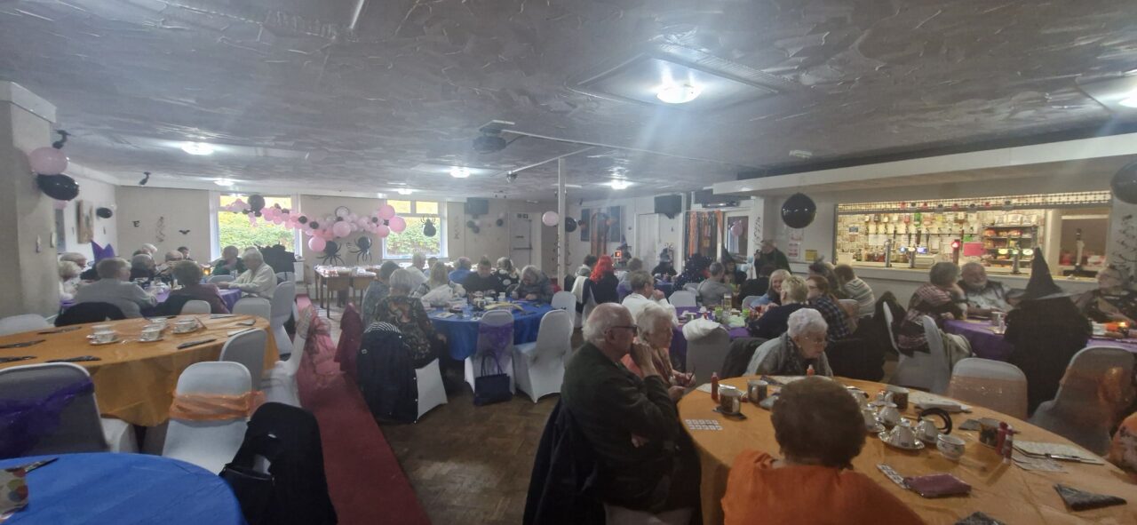 Delwedd o ystafell yn llawn pobl hŷn, pob un yn eistedd mewn grwpiau o amgylch byrddau. // Image of a room full of older people, all sat in groups around tables.