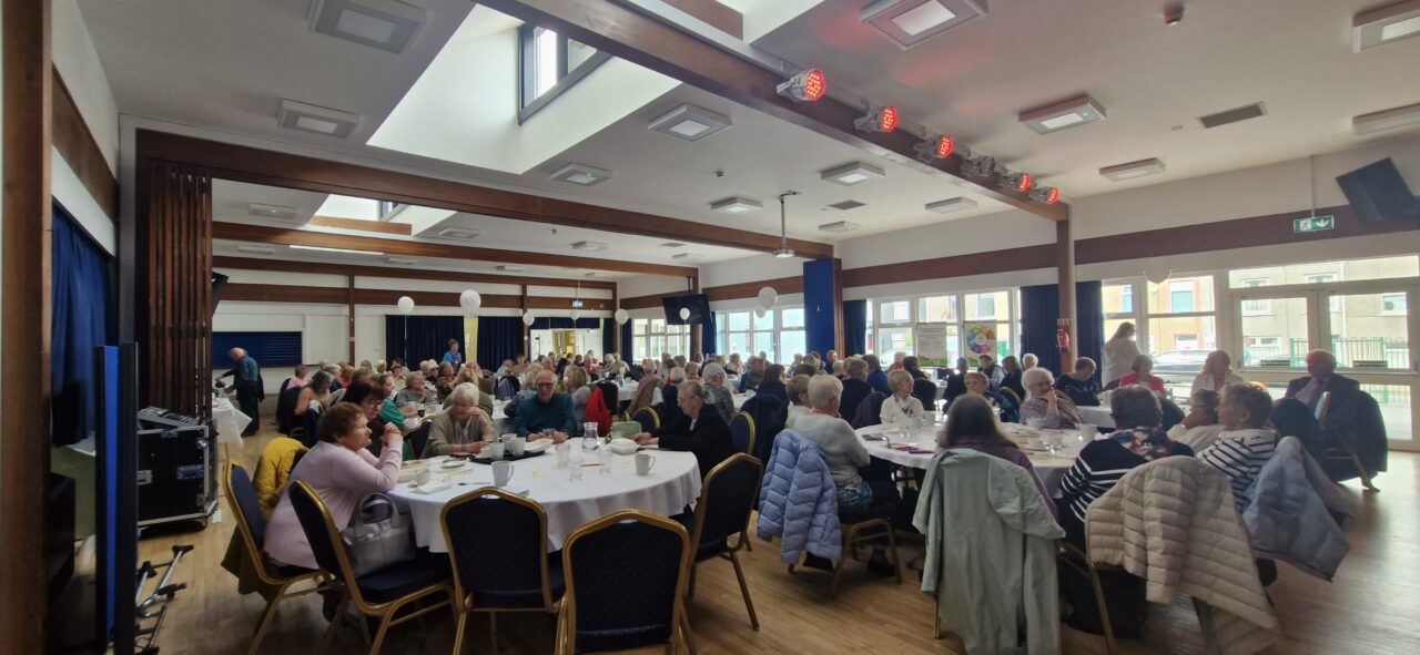 Delwedd o tua 120 o bobl hŷn yn eistedd o amgylch byrddau mewn neuadd fawr. // Image of roughly 120 older people sat around tables in a large hall.