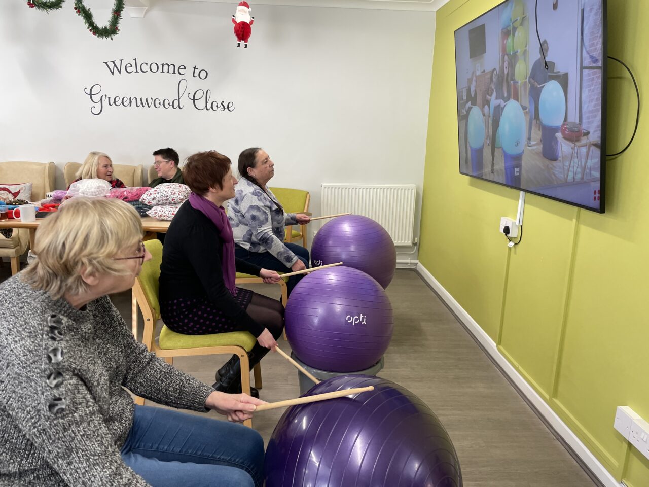 Comisiynydd Pobl Hŷn Cymru, Rhian Bowen-Davies (canol), yn rhoi cynnig ar ddrymio cardio gyda ffyn drymiau, pêl ioga a bwced. Mae dau berson hŷn ar y naill ochr a'r llall iddi hefyd yn ymuno. Mae'r tri yn eistedd ar gadeiriau y tu mewn i neuadd gymunedol. // Older People's Commissioner for Wales, Rhian Bowen-Davies (centre), having a go at cardio drumming with drum sticks, a yoga ball and a bucket. Two older people either side of her are also joining in. All three are sat on chairs inside a community hall.