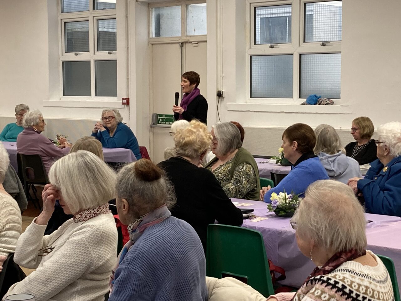 Comisiynydd Pobl Hŷn Cymru, Rhian Bowen-Davies, yn sefyll i fyny, gyda meicroffon yn ei llaw, yn rhoi araith i ystafell yn llawn pobl hŷn, sy'n eistedd o amgylch byrddau hir, yn ei gwylio. // Older People's Commissioner for Wales, Rhian Bowen-Davies, standing up, with a microphone in hand, giving a speech to a room full of older people, who are sat around long tables, watching her.