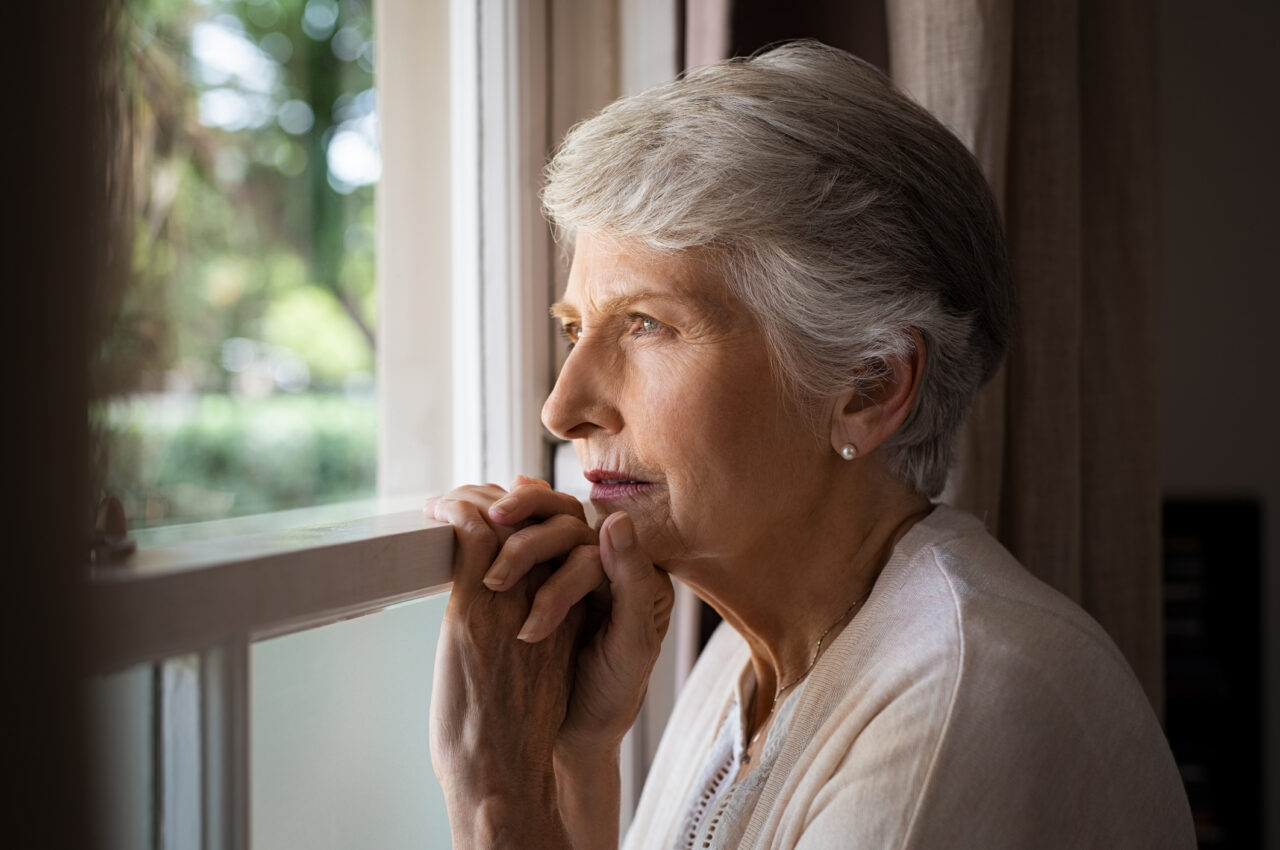 Menyw hŷn yn gwisgo crys pinc, yn sefyll i fyny ac yn edrych allan o'i ffenestr. // Older woman wearing a pink shirt, standing up and looking out of her window.
