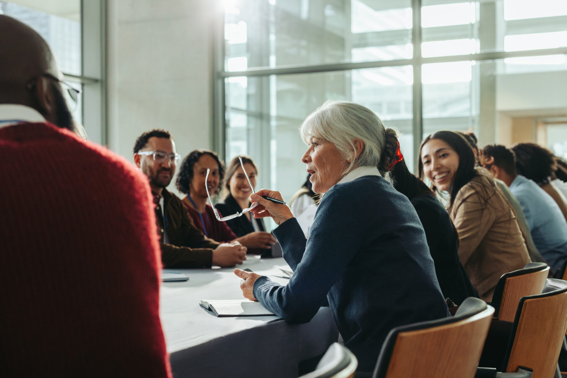 Eisteddodd grŵp o oedolion o wahanol oedrannau o amgylch bwrdd cynadledda, yn trafod. // A group of adults of various ages sat around a conference table, in discussion.