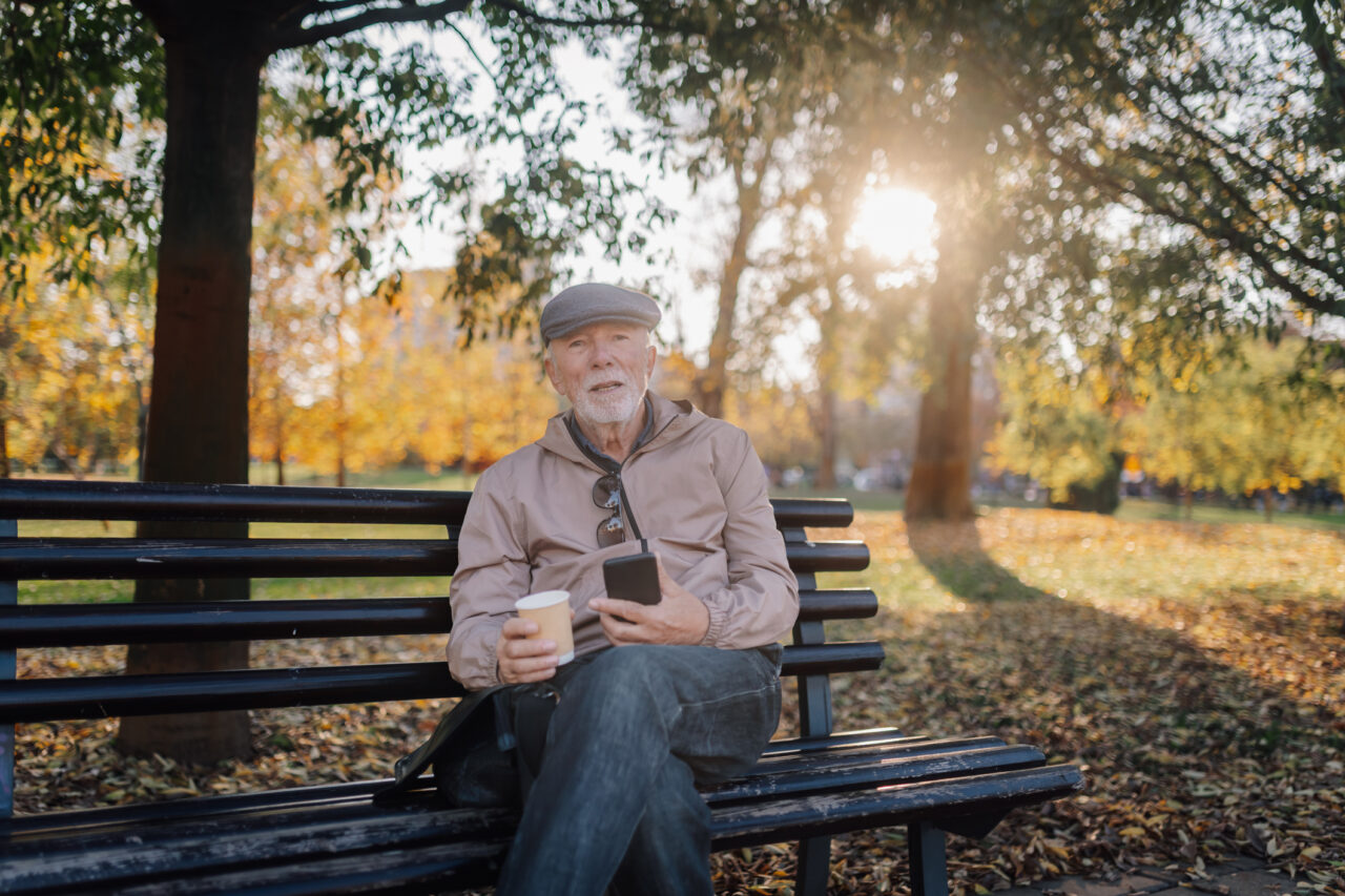 Dyn hŷn yn eistedd ar fainc parc, yn dal ffôn clyfar a choffi, yn mwynhau diwrnod tawel yn yr hydref yn yr awyr agored // Elder man sitting on a park bench, holding a smartphone and coffee, enjoying a tranquil autumn day outdoors