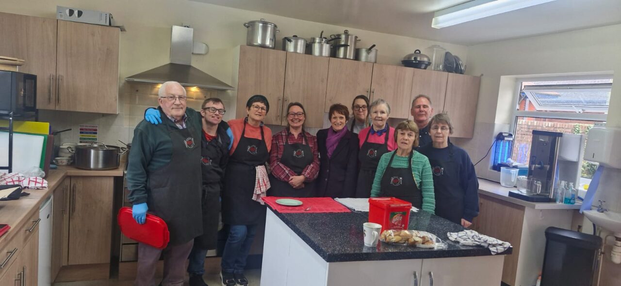 Comisiynydd Pobl Hŷn Cymru, Rhian Bowen-Davies (canol-dde), mewn cegin ochr yn ochr â gwirfoddolwyr yng Nghaffi Cymunedol Newtown. // Older People's Commissioner for Wales, Rhian Bowen-Davies (centre-right), pictured in a kitchen alongside volunteers at Newtown Community Cafe.