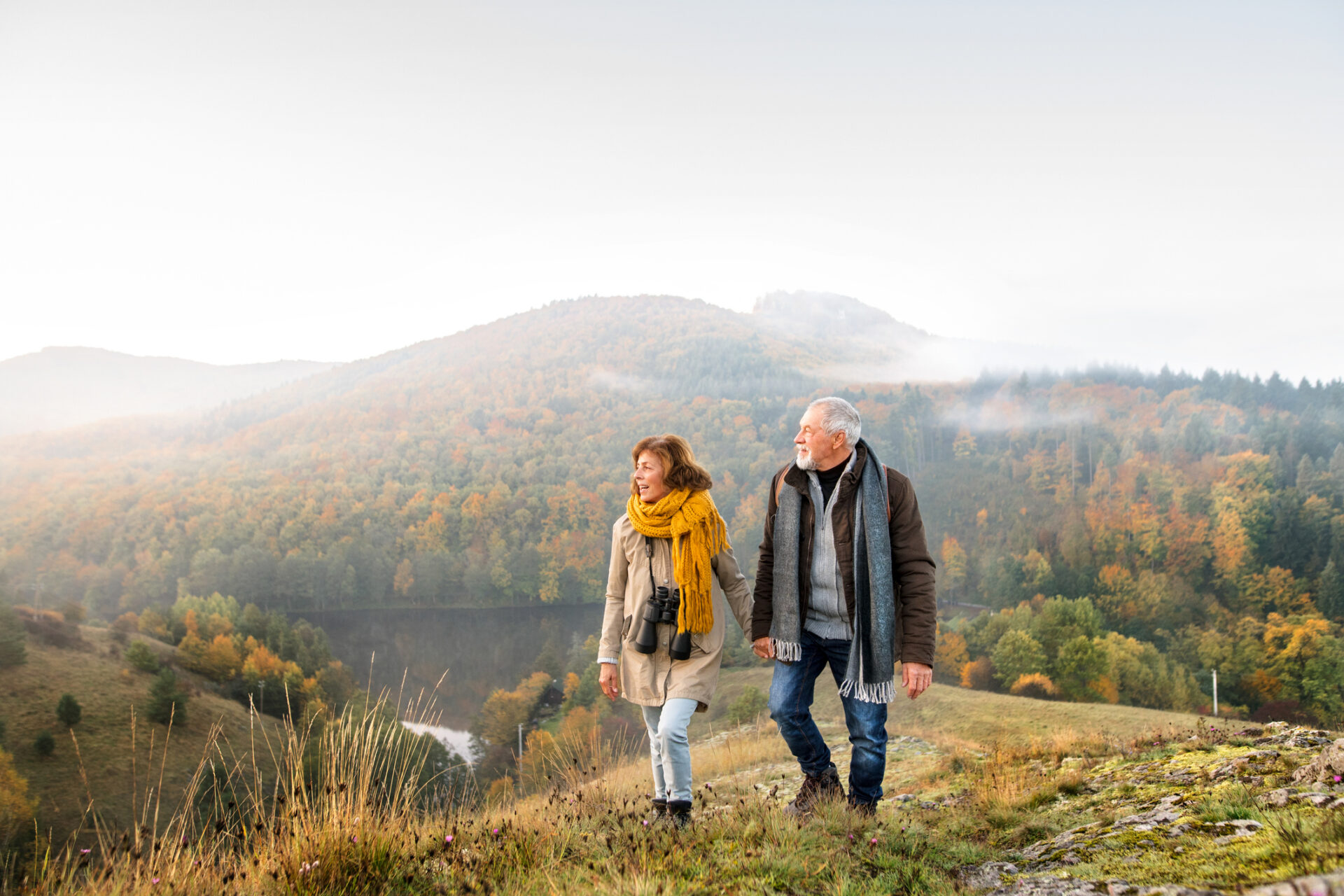 Cwpl hŷn yn cerdded law yn llaw dros fryn, yn edrych dros lyn. / Older couple walking hand in hand over a hill, overlooking a lake.