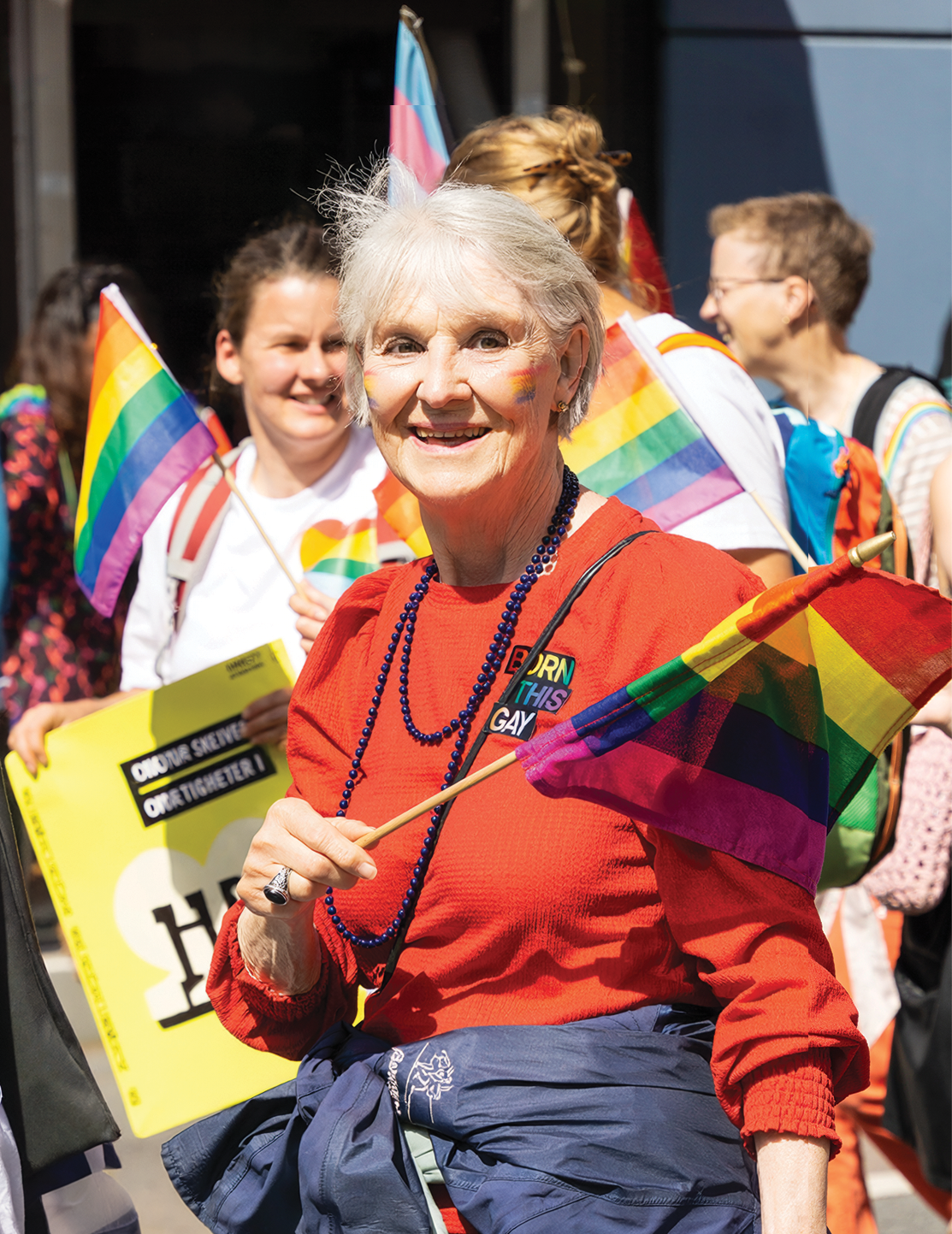 Photograph of a participant at a Pride event holding a rainbow flag. The participant is wearing a bright red top, blue bead necklaces and a jacket tied around the waist. Several other participants are visible in the background, some holding rainbow and transgender pride flags. One person is carrying a yellow sign. The image is brightly lit by sunlight and features prominent rainbow colours throughout.