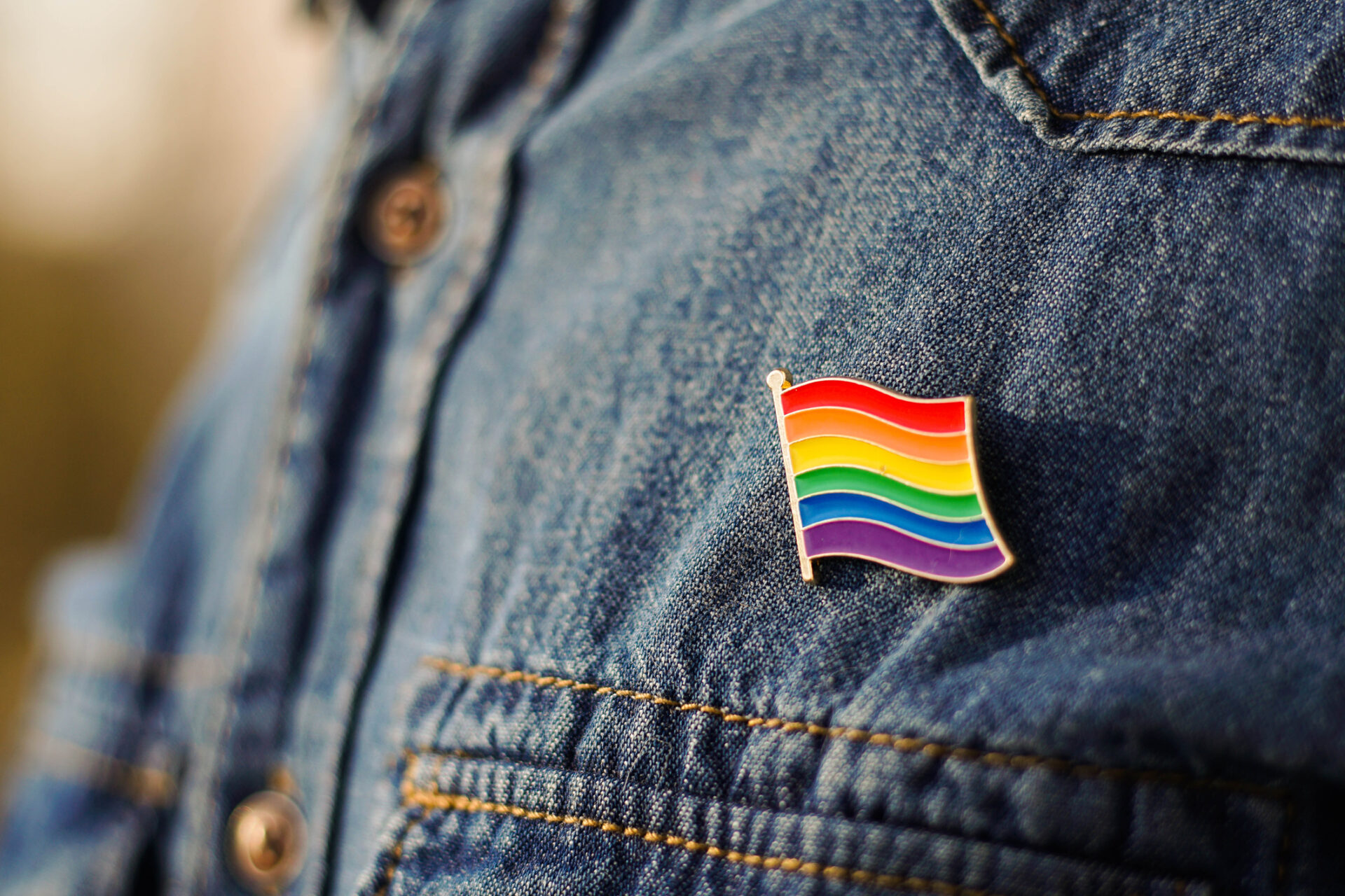 Close-up of a denim jacket with a rainbow Pride flag pin attached to the chest pocket. The brightly coloured pin stands out against the blue denim, with a softly blurred background.